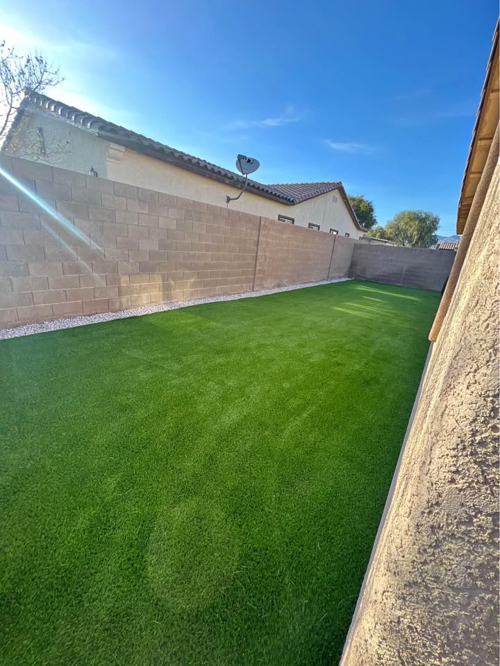 A backyard with bright green artificial grass, bordered by tan brick walls and houses on either side, under a clear blue sky with sunlight casting shadows on the ground.