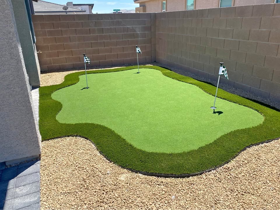 A backyard putting green with three holes and flags, surrounded by artificial turf and bordered by gravel and stone walls in a small enclosed outdoor space.