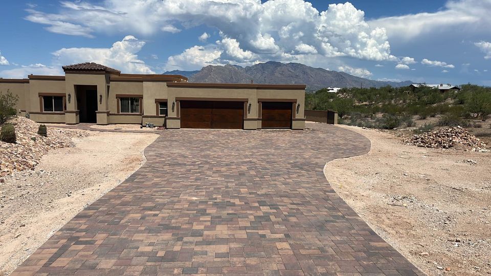 A modern, single-story house in Carefree AZ features a long, wide, patterned pavers installation driveway in a desert landscape. Mountains and a partly cloudy sky are visible in the background, with sparse greenery surrounding the home.