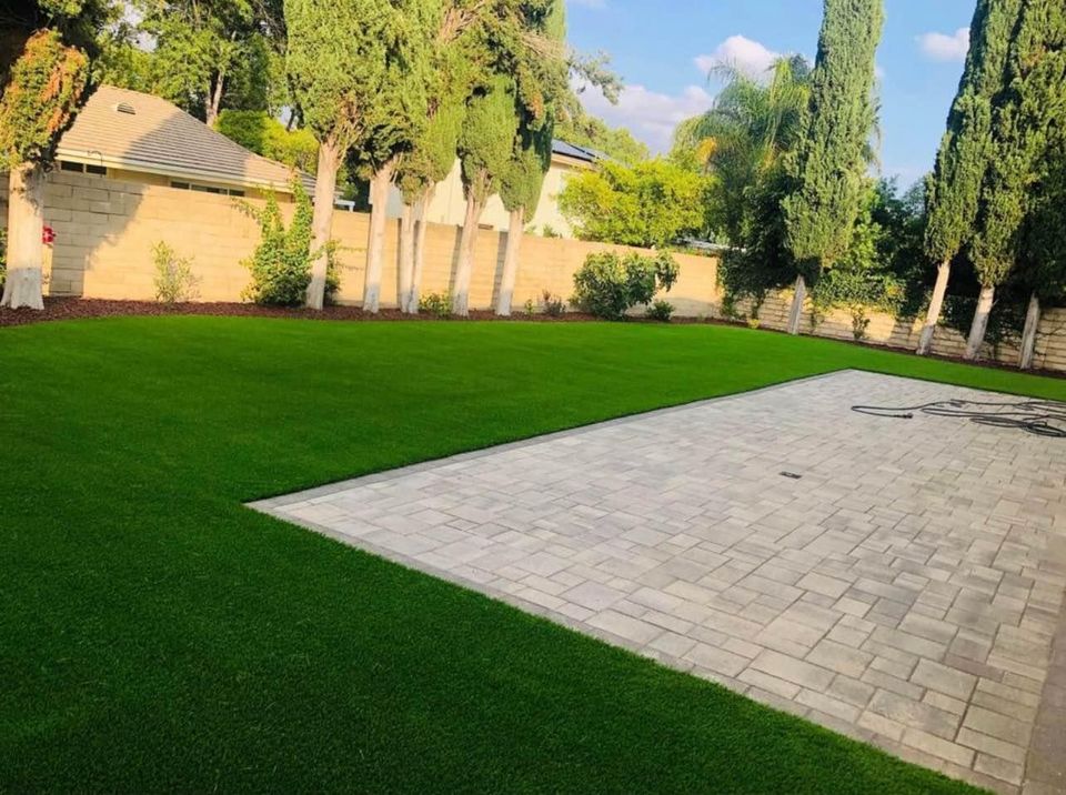 A backyard with a large area of lush green grass—featuring artificial grass for easy maintenance in Carefree, AZ—plus a paved patio and tall trees lining a stone wall fence under a partly cloudy sky.