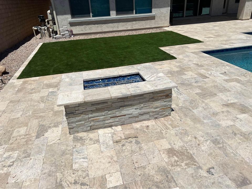 A rectangular stone fire pit sits on a light-colored tile patio near a small patch of lush artificial grass, offering low-maintenance beauty for any backyard in Carefree, AZ, with a sparkling swimming pool nearby.